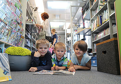 Drei Kinder liegen auf dem Teppich einer Fahrbücherei und betrachten Bücher.