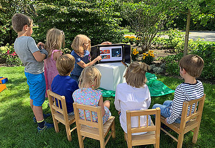 Eine Frau sitzt gemeinsam mit einer Gruppe von Kindern an einem Tisch im Garten und zeigt etwas in einem Buch.